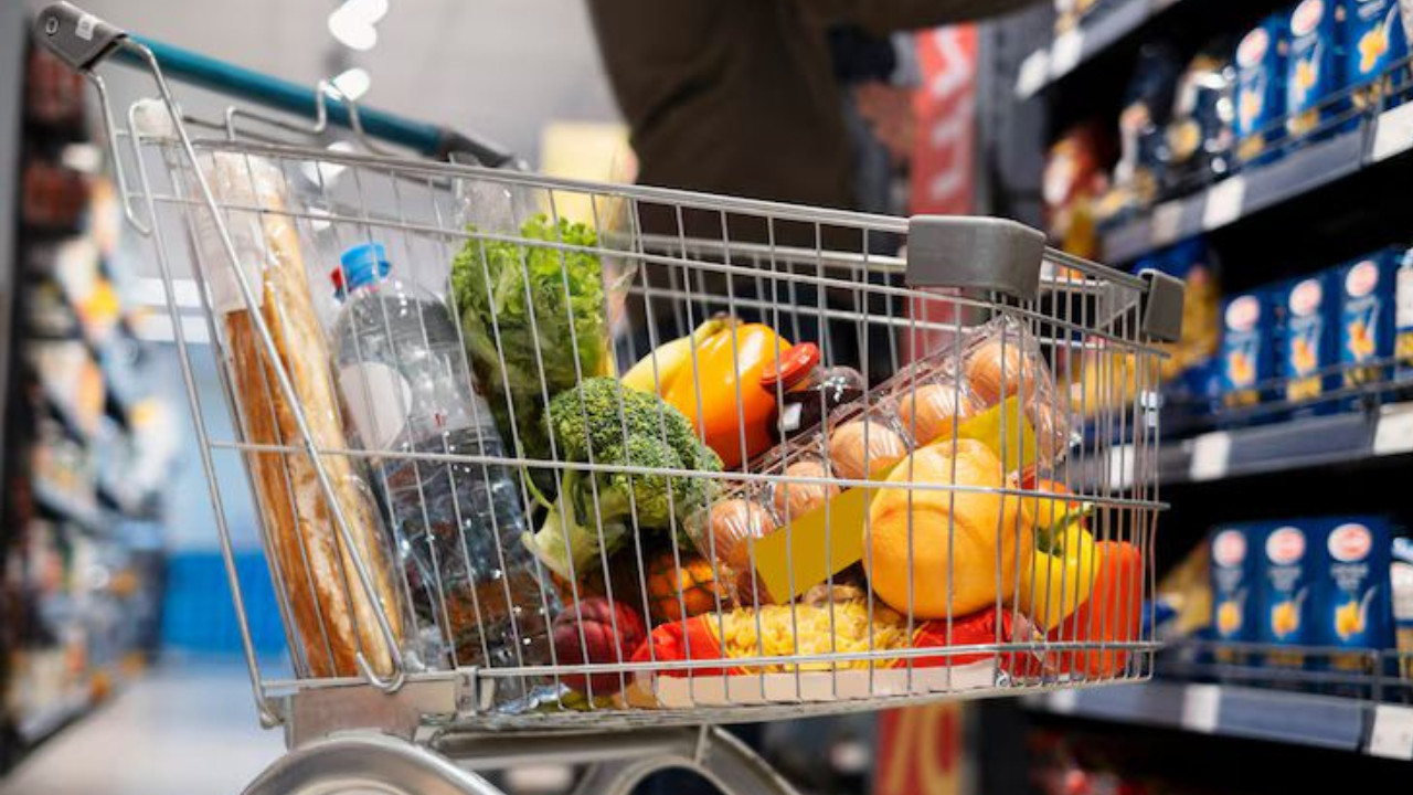 Carrinho de supermercado cheio de alimentos diversos como ovos, frutas, vegetais frescos, pão, água e produtos industrializados em meio a um corredor com prateleiras abastecidas e uma pessoa fazendo cotações. A imagem remete à importância de fazer boas cotações de preços antes das compras, especialmente em épocas de alta demanda no varejo.