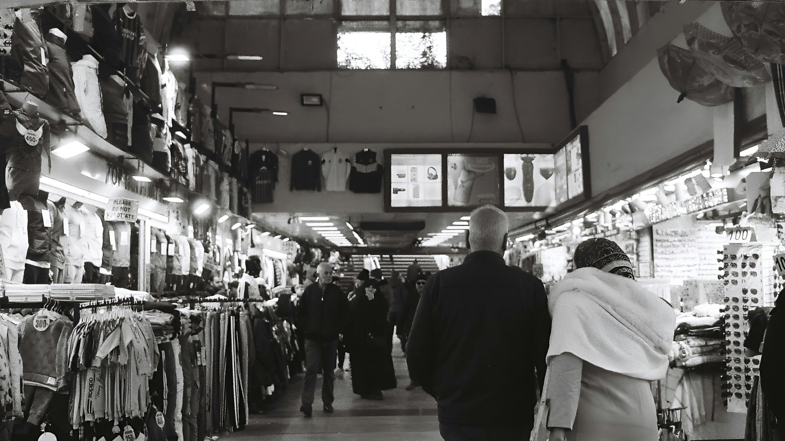 Corredor interno de um mercado popular coberto, com diversas barracas vendendo roupas, acessórios e itens variados. Um casal caminha de costas em direção ao fundo do corredor, cercado por vitrines iluminadas e manequins. Ao fundo, várias pessoas circulam, criando um ambiente movimentado e informal. Apesar da agitação pública, a interação entre o casal transmite um momento private e íntimo em meio à multidão.