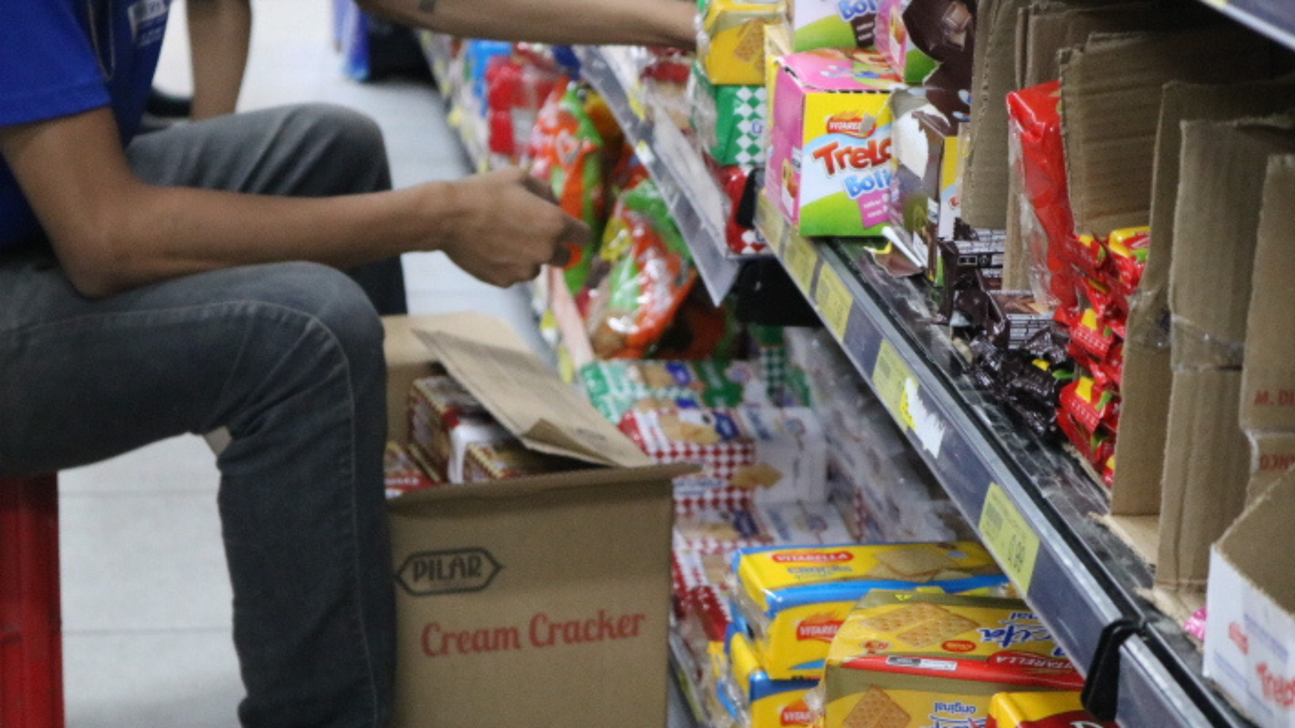 Funcionário organiza prateleiras de biscoitos e snacks em um supermercado, enquanto produtos são repostos em meio a caixas abertas. A inflação impacta diretamente o preço desses itens básicos, afetando o consumo diário das famílias